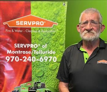 Man with a white beard and glasses in a green servpro labeled golf shirt next to a colorful banner